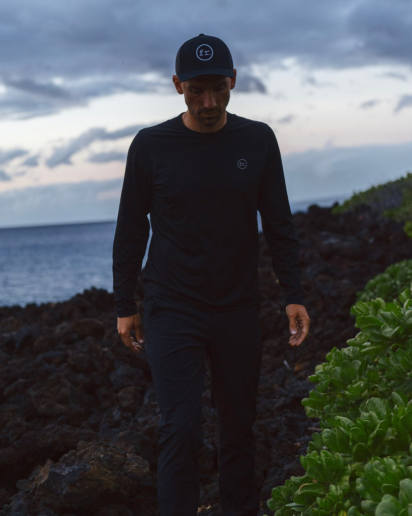 Man walking on lava rocks wearing Foreign Rider Co hat, t-shirt and Performance pants