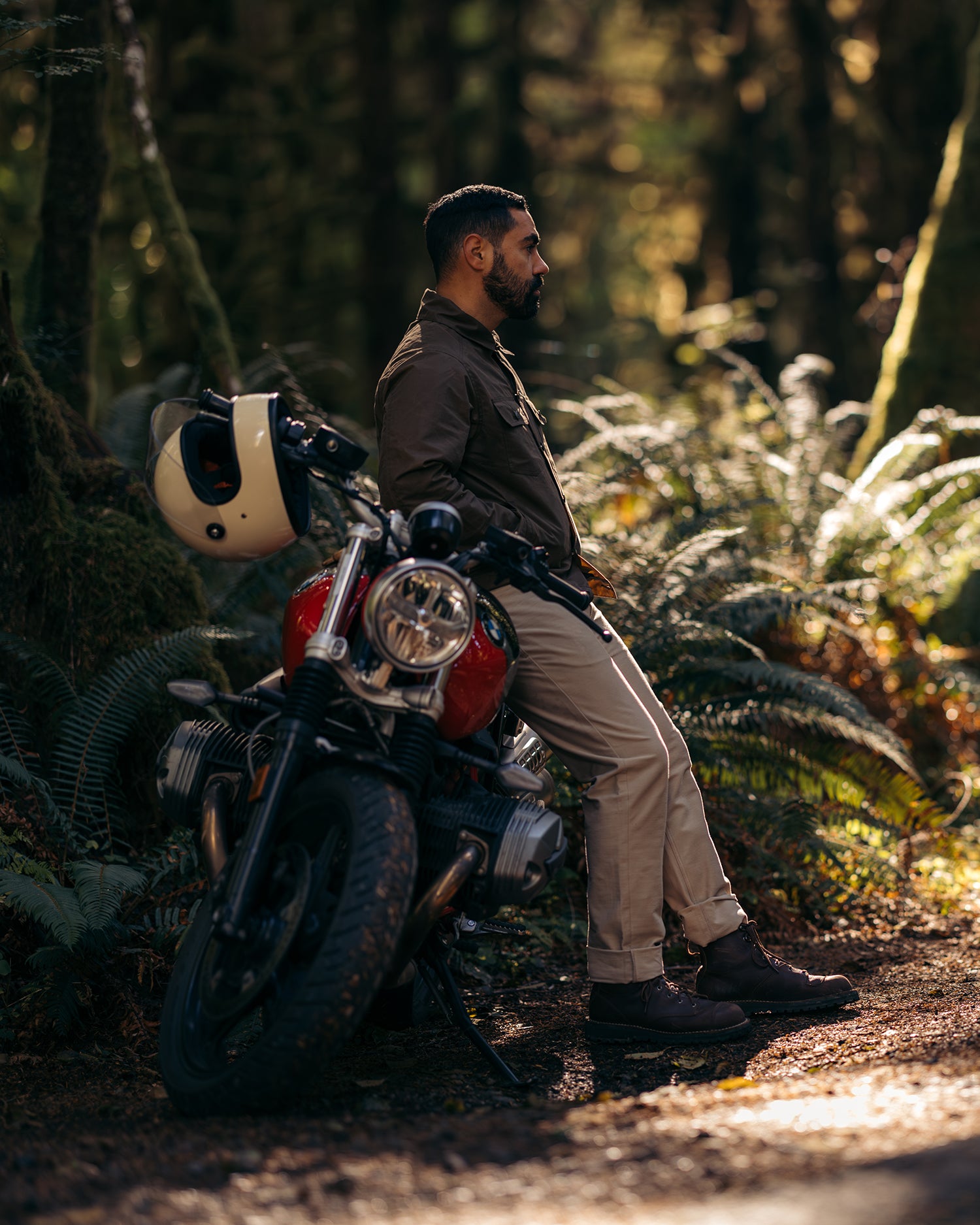 Man sitting on the side of parked motorbike alongside a forested road wearing Foreign Rider clothing
