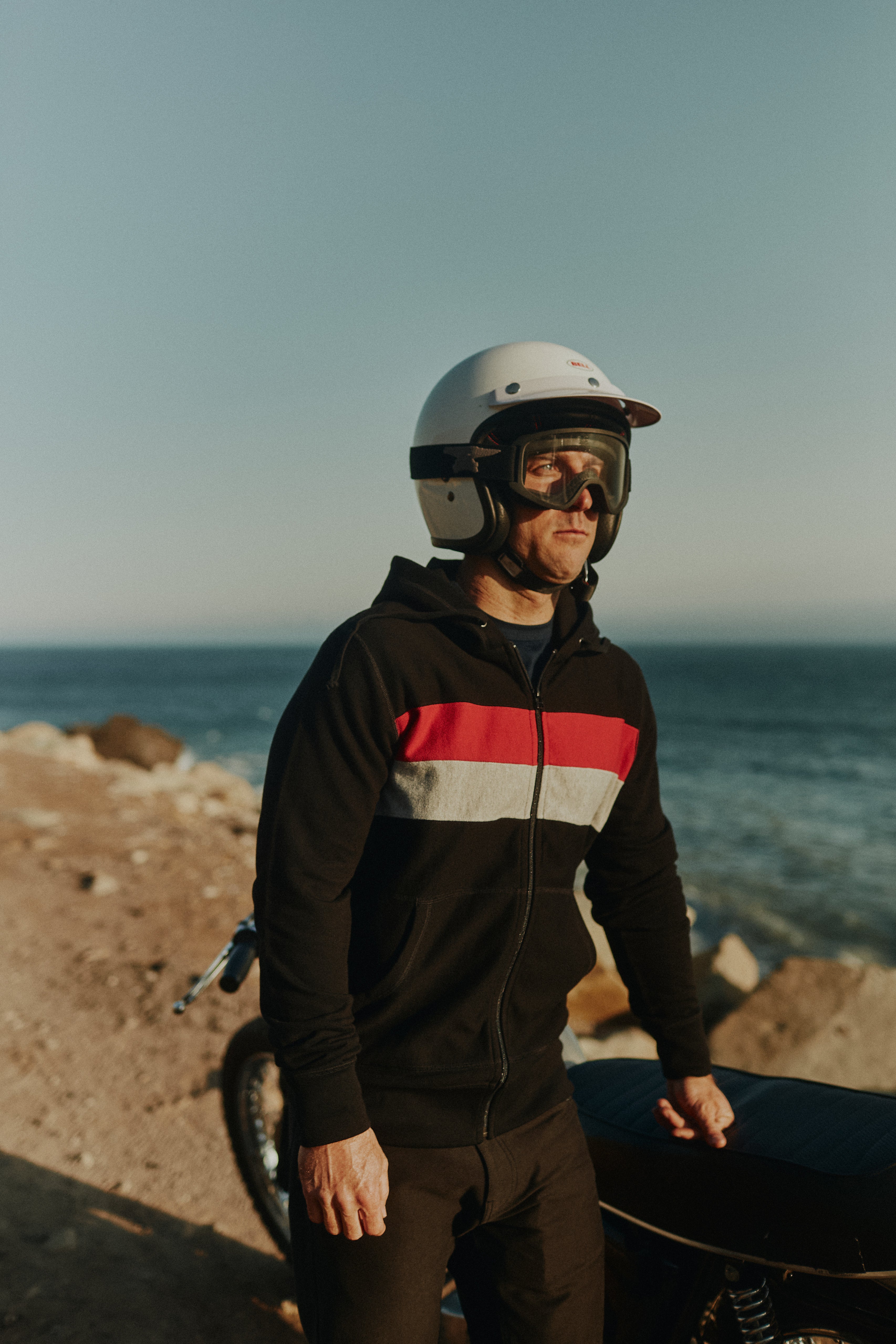 Man on a dirt road standing next to a vintage motorbike overlooking the Pacific ocean wearing a helmet and the Foreign Rider Striped Full Zip Hooded Sweatshirt