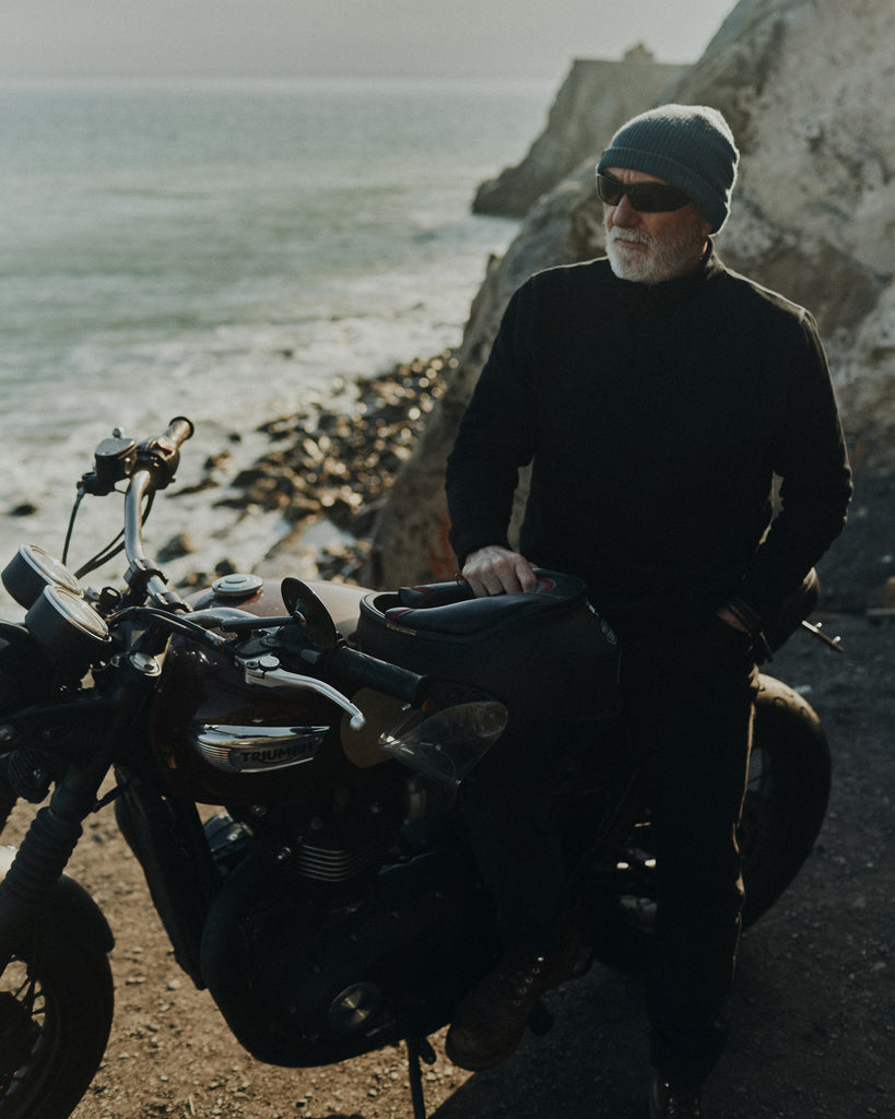 Ralph Dunning sitting on a vintage Triumph motorbike overlooking the Pacific ocean wearing the Foreign Rider Quarter Zip Sweatshirt
