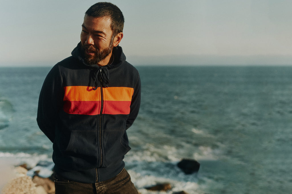 Man standing on a cliff overlooking the Pacific ocean wearing the Foreign Rider Striped Full Zip Hooded Sweatshirt