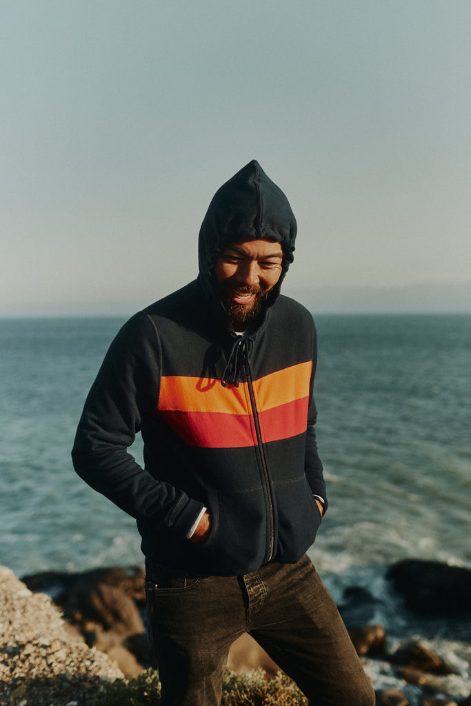 Man standing on a cliff overlooking the Pacific Ocean wearing the Foreign Rider Striped Full Zip Hooded Sweatshirt with the hood up