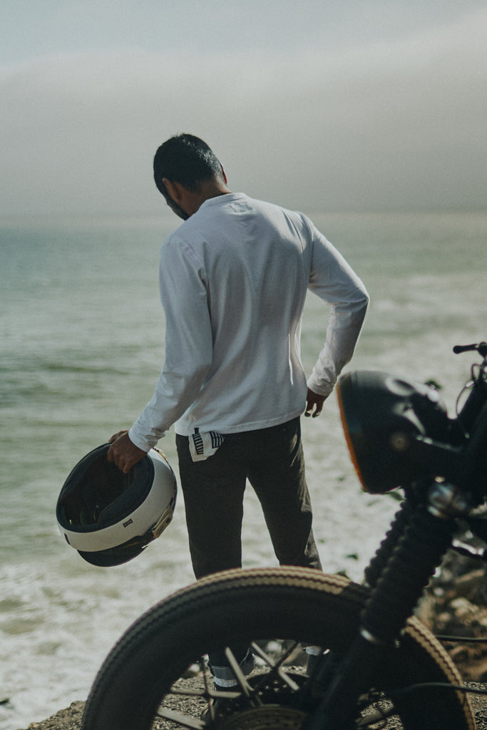 Man looking down at the Pacific ocean from a cliff holding a motorbike helmet wearing the Foreign Rider Supima Long Sleeve T-shirt