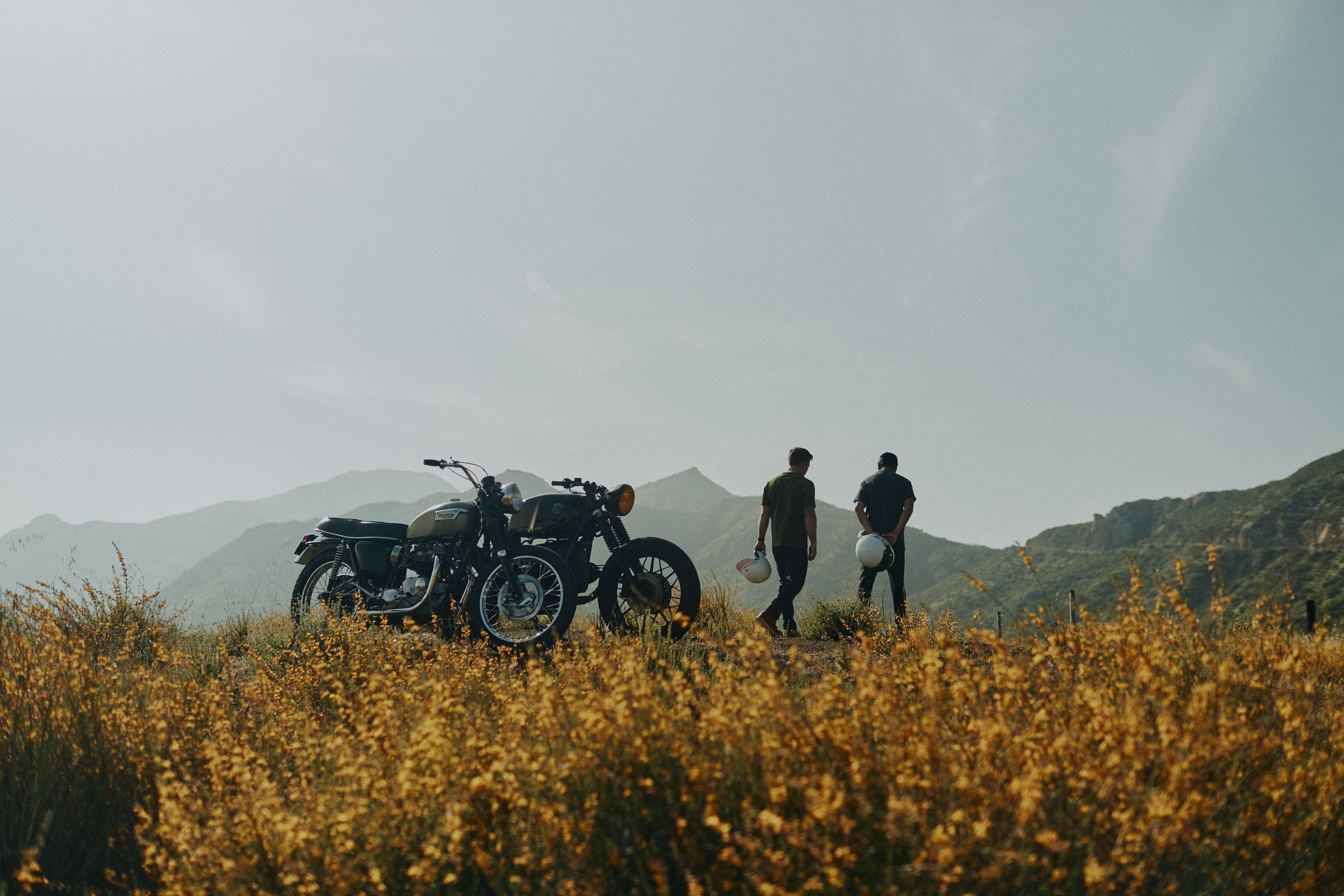 Low landscape view of yellow flowers, mountains and skyline with 2 men walking away from vintage motorbikes wearing the Foreign Rider Supima Short Sleeve T-Shirt