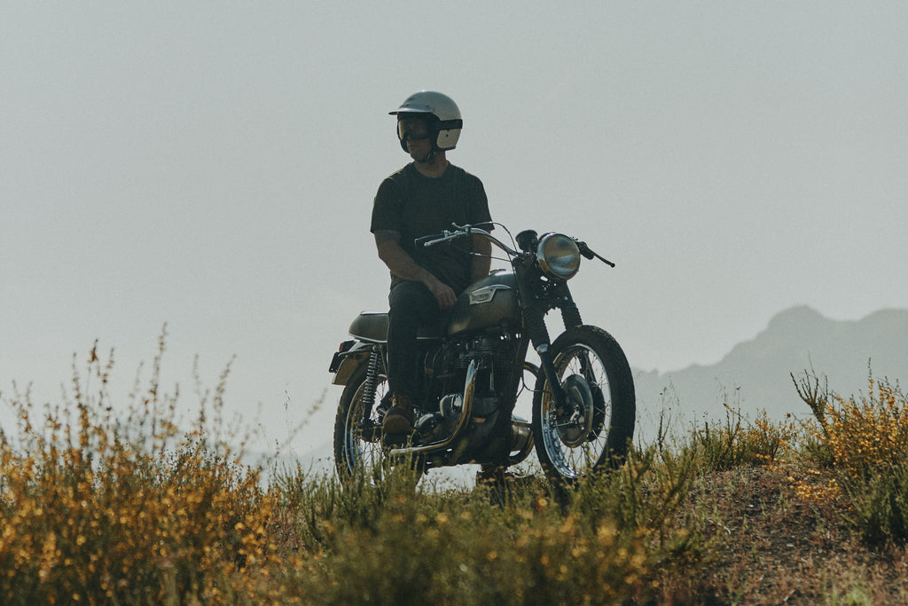 Low landscape view of grass, yellow flowers, mountains and blue sky with a man sitting on a vintage Triumph motorbike wearing the Foreign Rider Supima Short Sleeve T-shirt