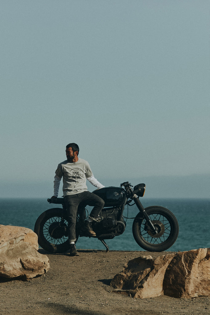 Man on a gravel road with rocks sitting on an idle vintage motorbike with a blue sky and Pacific ocean in the background wearing Foreign Rider Tiger Graphic T-Shirt