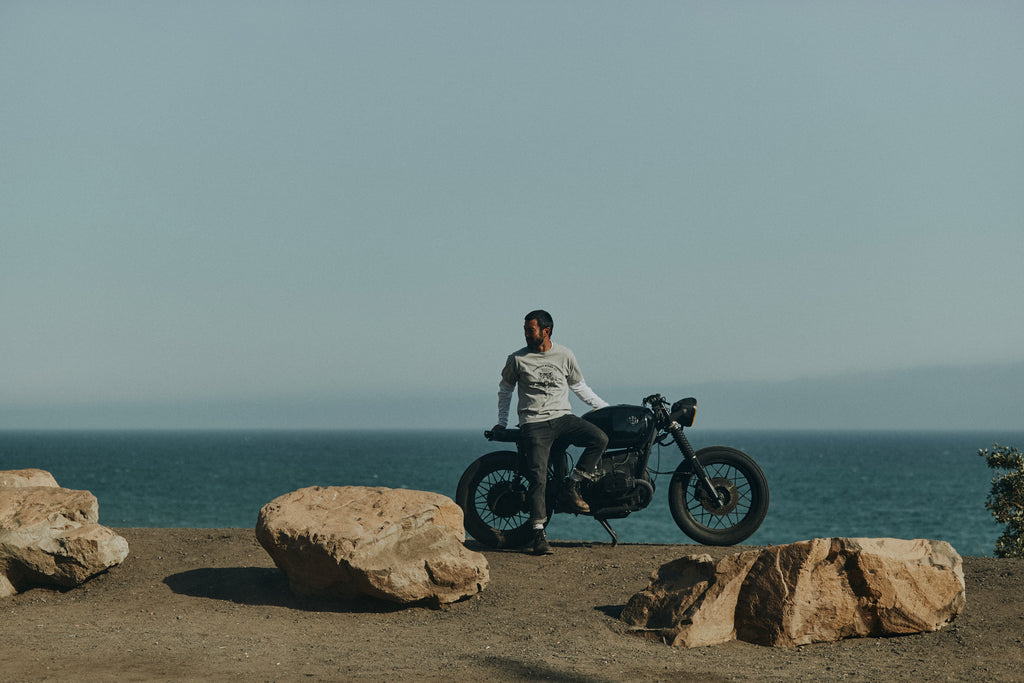 Man on a gravel road with rocks sitting on an idle vintage motorbike with a blue sky and Pacific ocean in the background wearing Foreign Rider Tiger Graphic T-Shirt