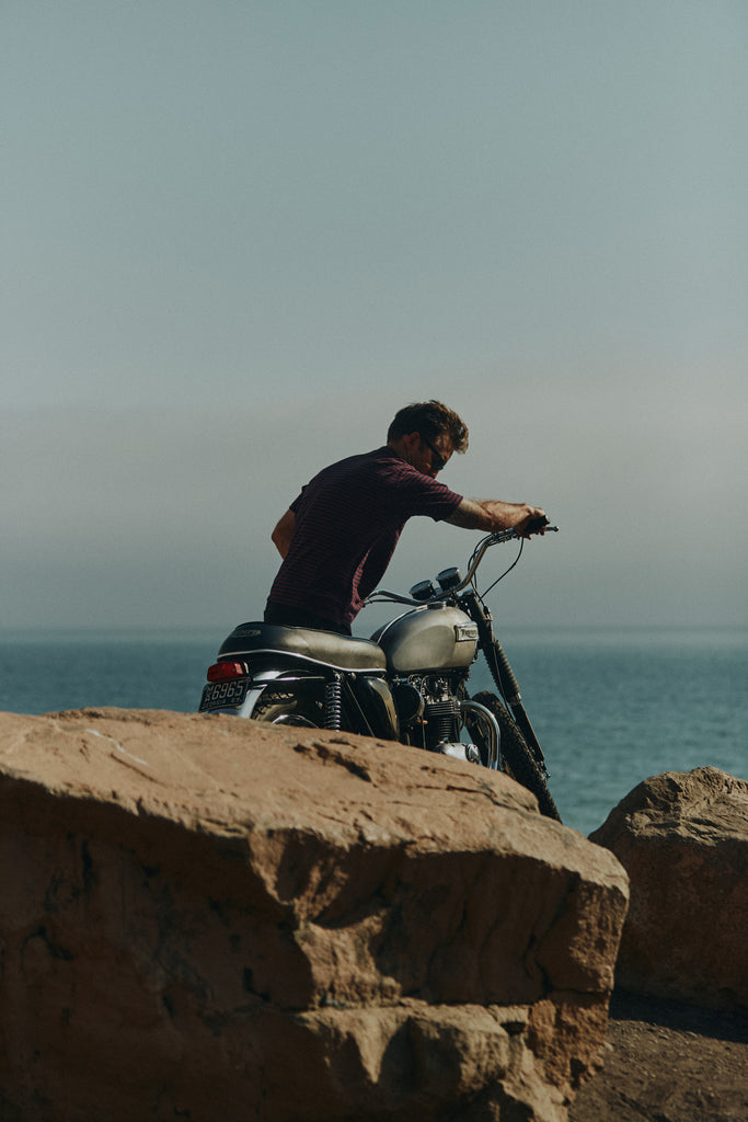Man moving his motorbike on a gravel and rocky path that overlooks the Pacific ocean wearing the Foreign Rider Vintage Stripe T-Shirt 