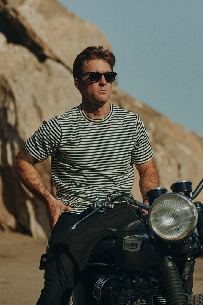 Man on gravel with rocks sitting on idle Triumph motorbike wearing the Foreign Rider Vintage Stripe T-Shirt