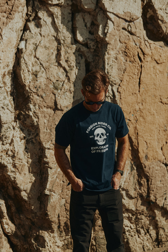 Man standing in front of a rock wall looking down wearing the Foreign Rider Skull Graphic T-shirt