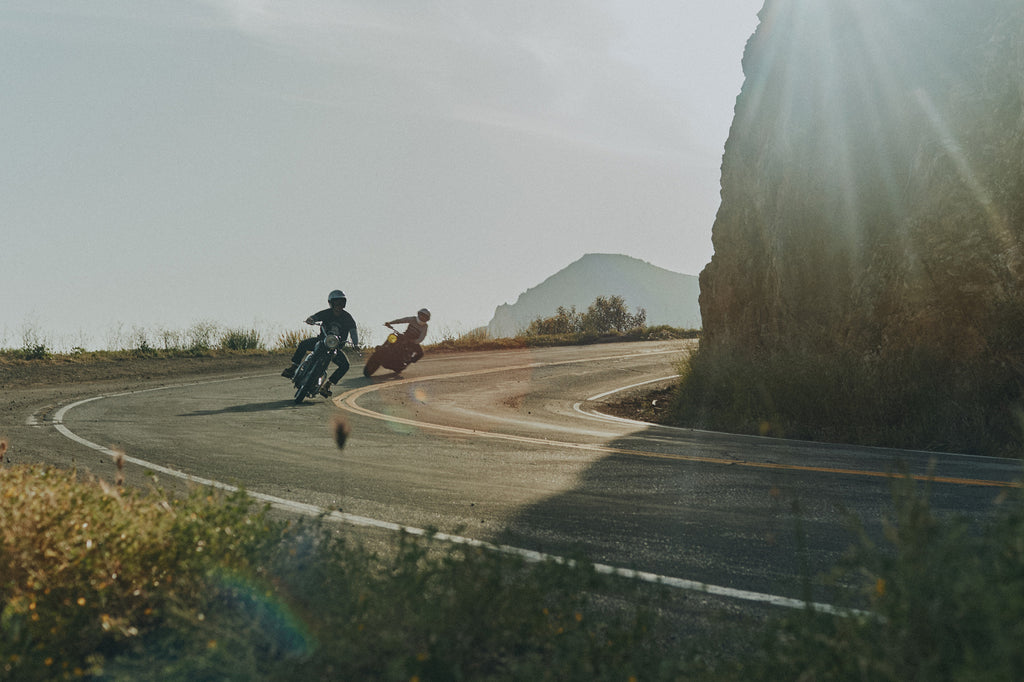 Two men on vintage motorcycles rounding a Malibu cliffside road