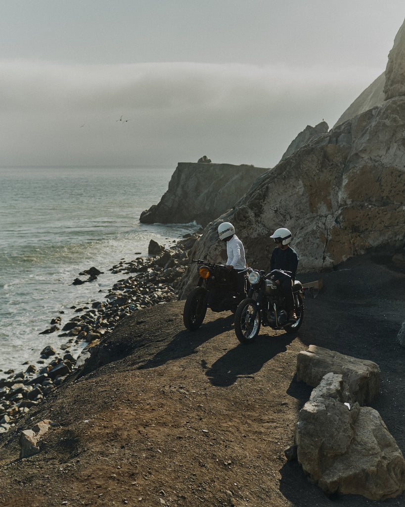 Two men sitting on their idle vintage motorbikes overlooking the Pacific ocean from a Malibu cliffside