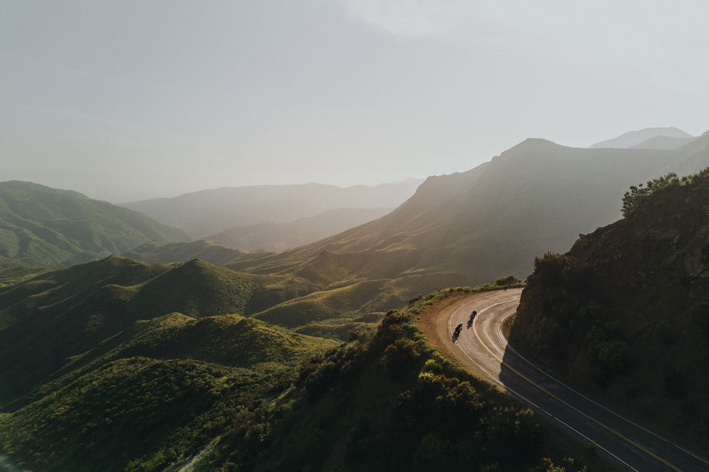 Landscape view of green Santa Monica mountains and two men rounding a corner on vintage motorbikes