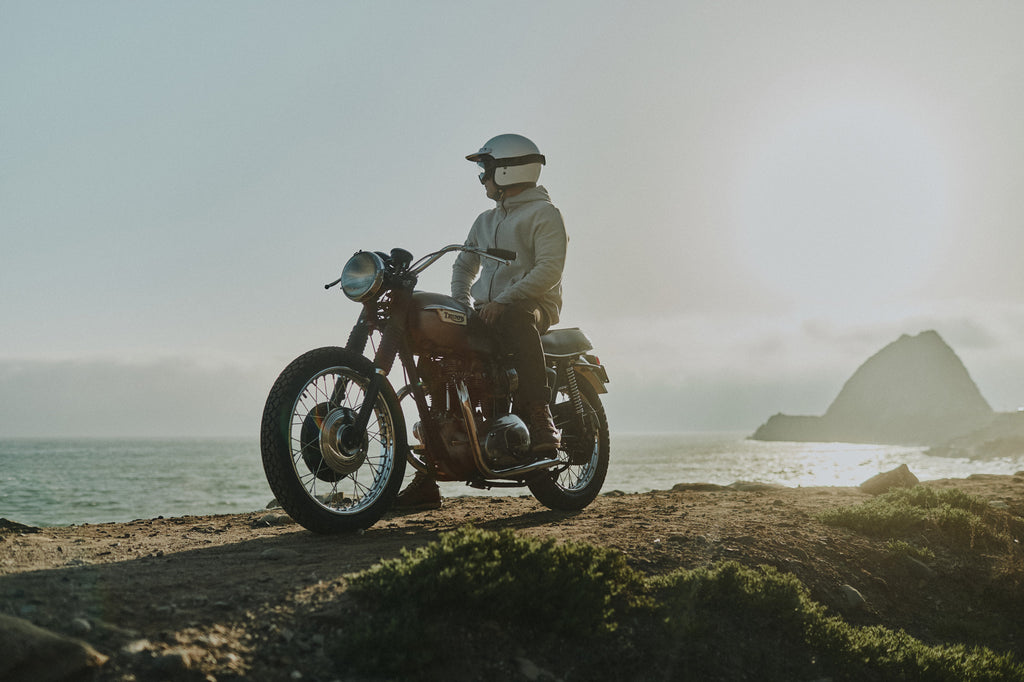 Man cliffside sitting on idle Triumph motorbike overlooking the Pacific ocean wearing Foreign Rider clothing