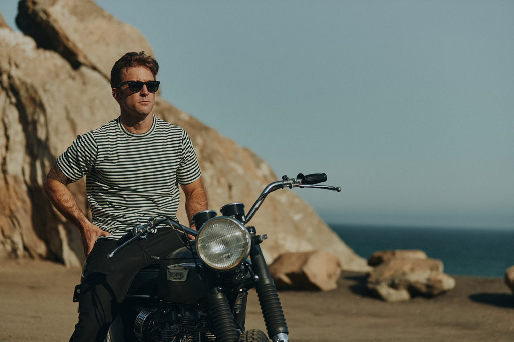 Man cliffside over the Pacific ocean sitting on parked Triumph vintage motorbike wearing Foreign Rider clothing