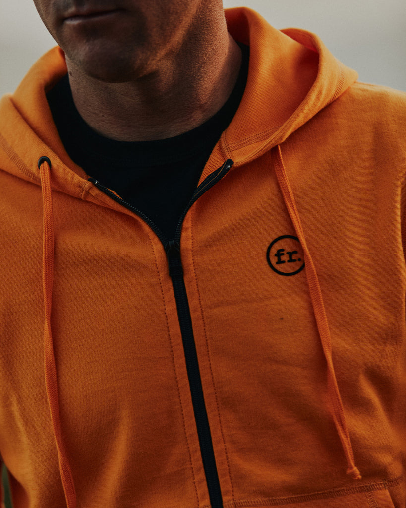 Chest detail shot of black YKK zipper and embroidered Foreign Logo on the orange Full Zip Hoodie