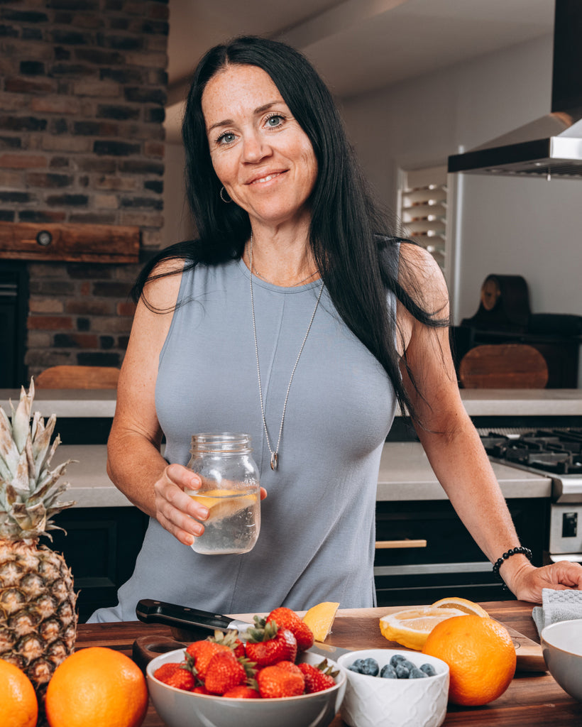 Shannon Kemp in a kitchen holding a glass of water in front of a bunch of fruit