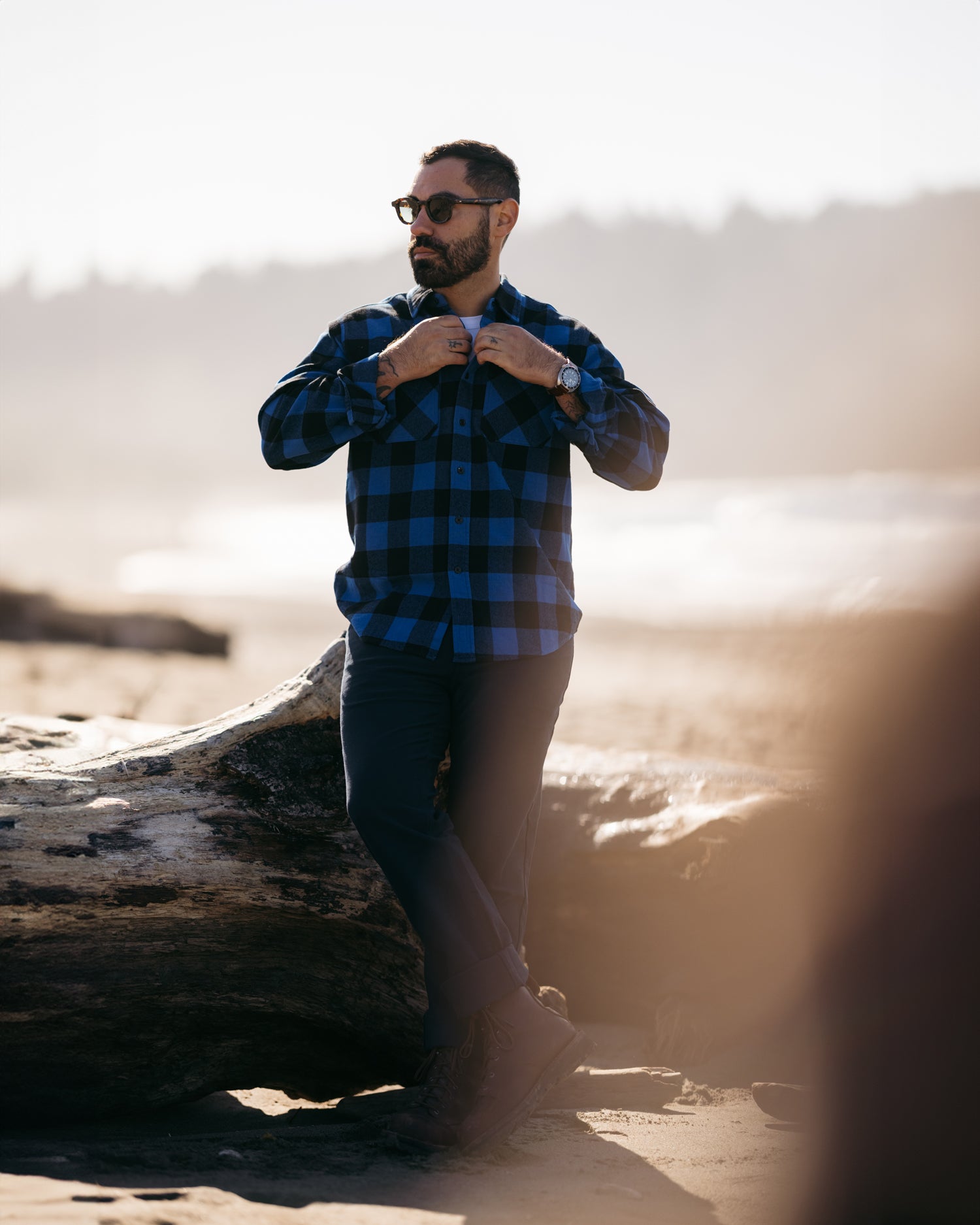 Man leaning on a stump on a beach unbuttoning blue Foreign Rider Co Flannel