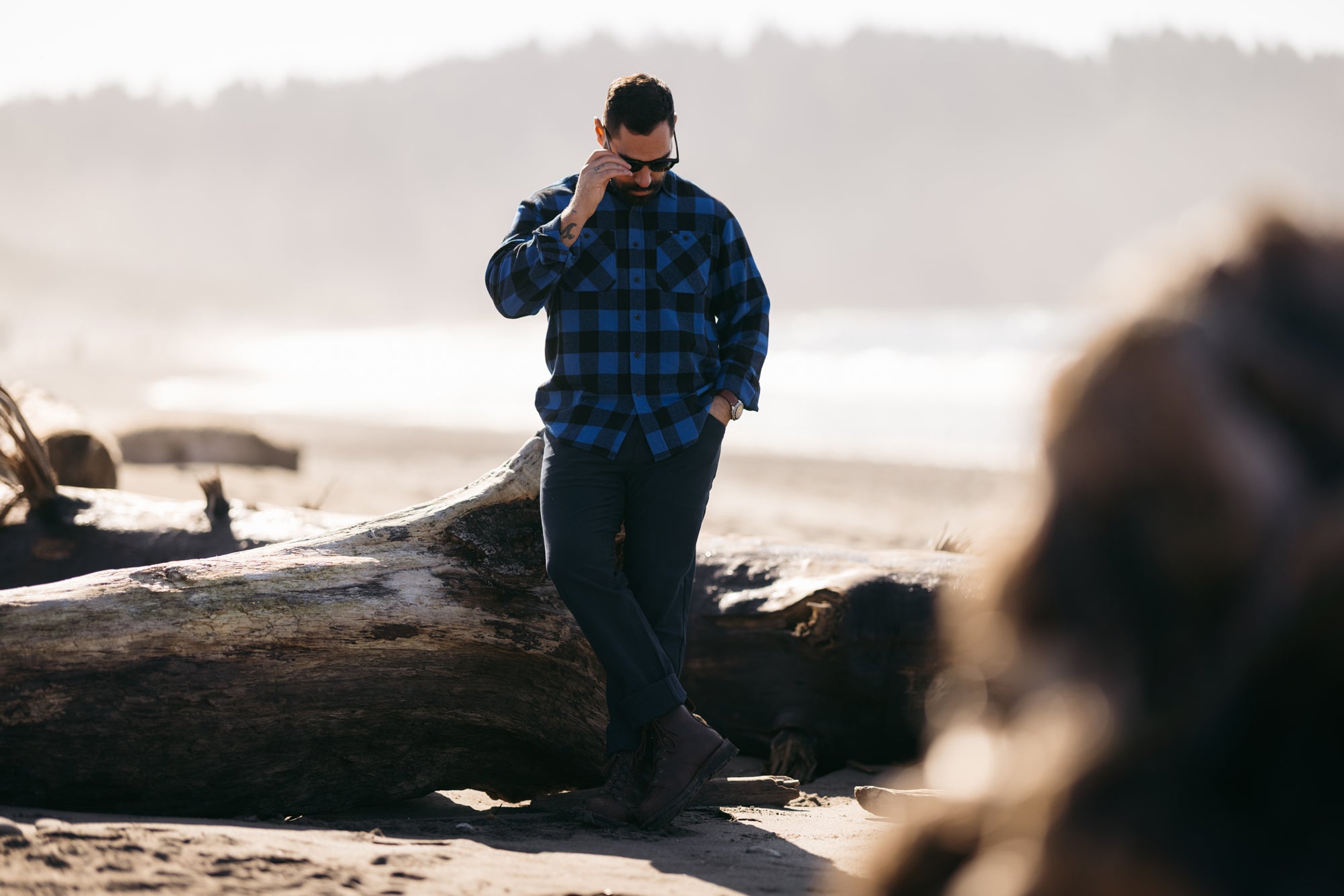 Man in sunglasses looking down leaning on a stump on a beach wearing Foreign Rider Co Blue Flannel