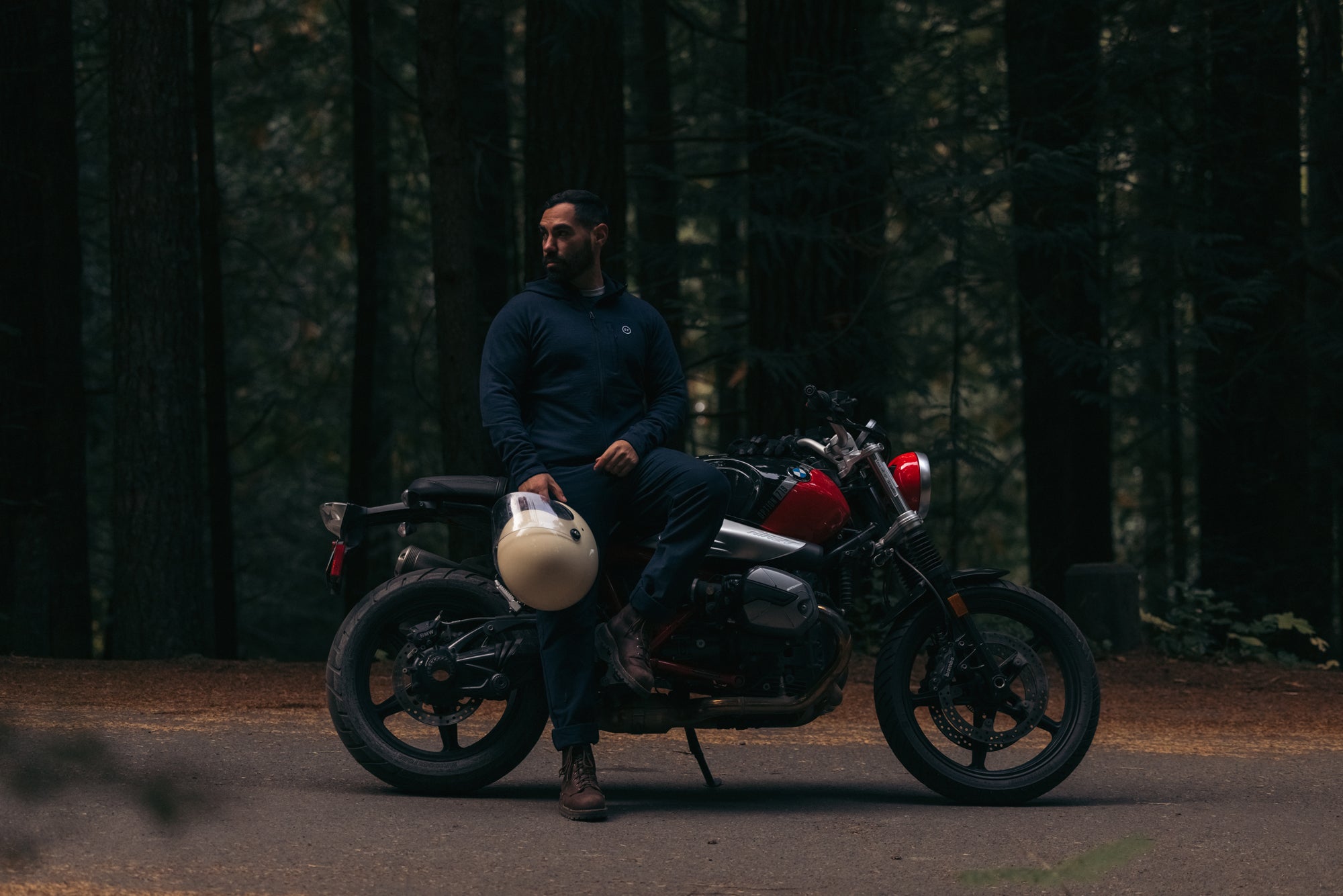 Man sitting on side of parked BMW Motorbike alongside forested road wearing Foreign Rider Co Merino Hooded Jacket