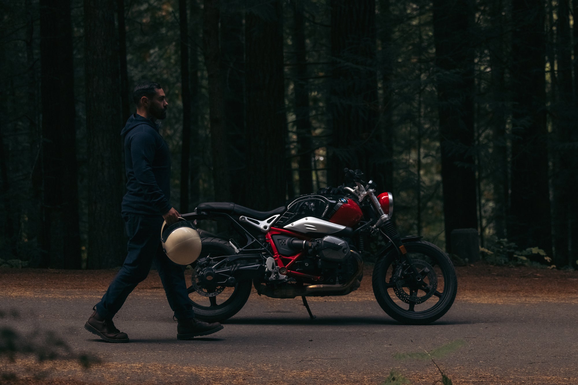 Side view of man walking alongside parked BMW motobike on forested road wearing Foreign Rider Co Merino Hooded Jacket