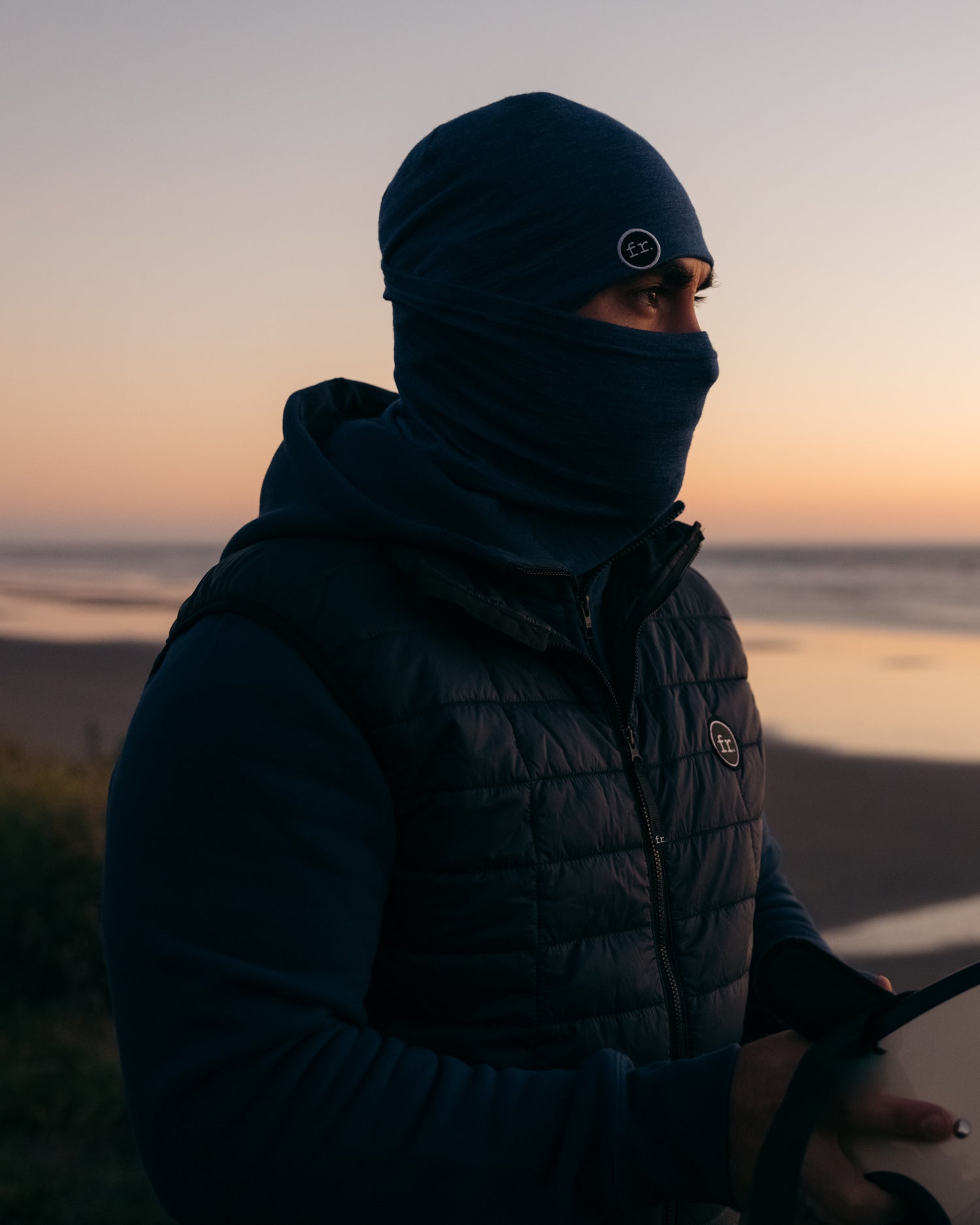 Side view of man standing on beach at sunset looking at ocean wearing Foreign Rider Co Merino Beanie and Necktube