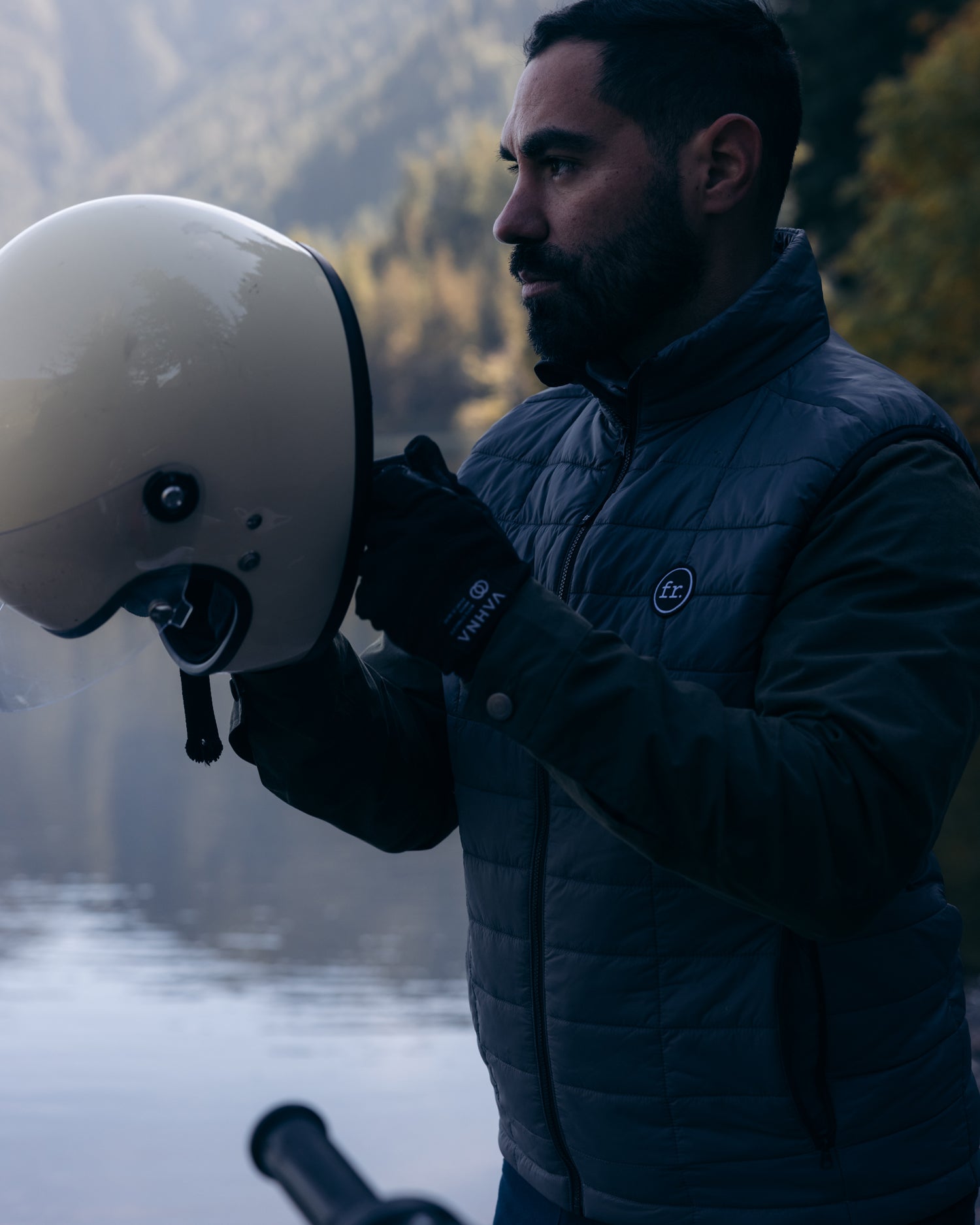 Man about to put on helmet in front of lake wearing Foreign Rider Co Insulated Vest