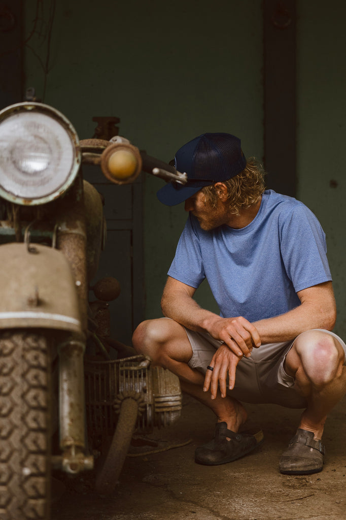 Eric Lagerstrom kneeling & inspecting a vintage rundown motorbike wearing the Foreign Rider Performance Short Sleeve T-Shirt
